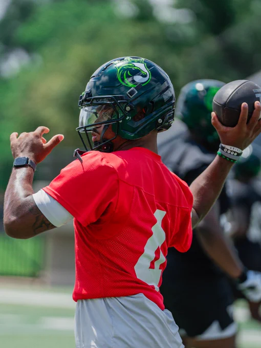 A football player wearing a red jersey and a black helmet with a team logo prepares to throw a football during practice. The scene exudes focus and determination.