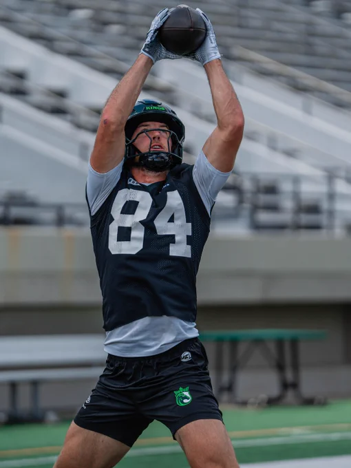 A football player in a practice session catches a ball overhead. The player wears a dark jersey with the number 84 and appears focused and determined.