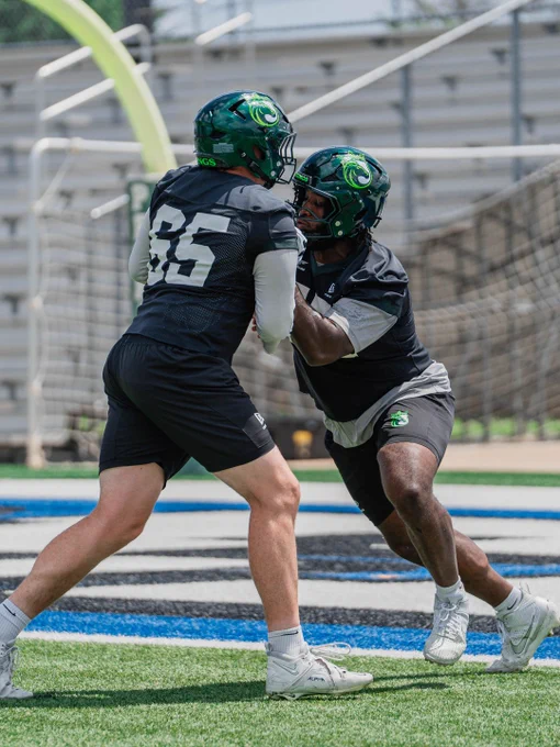 Two athletes engage in a football practice, wearing protective helmets and gear. The scene is set on an outdoor sports field.