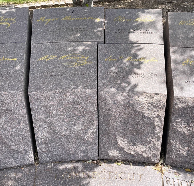 Four reddish gray stone blocks with names etched into the tops and gold signatures on them and the word "Connecticut" written at the base stand as a section of a low memorial wall.