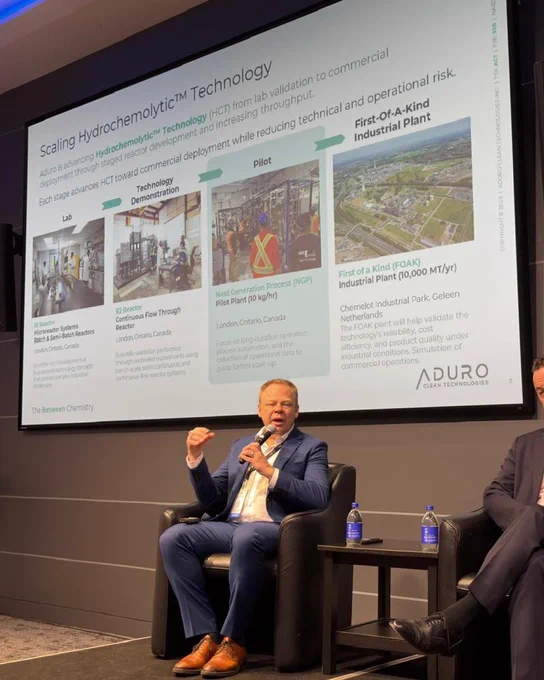 Abe Dyck sitting on stage speaking on a panel at the CCIC. A powerpoint slide in the background shows several stages of scaling Hydrochemolytic™ Technology.