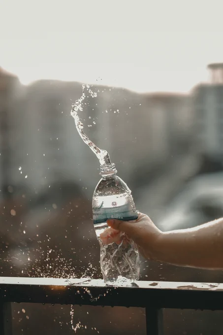 Hand squeezing a plastic water bottle, causing water to splash out in an arc against a blurred urban background.