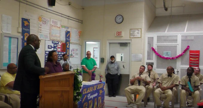 A man in a suit speaks at a podium to seated men in beige uniforms inside a classroom-like room with posters and a clock on the wall.