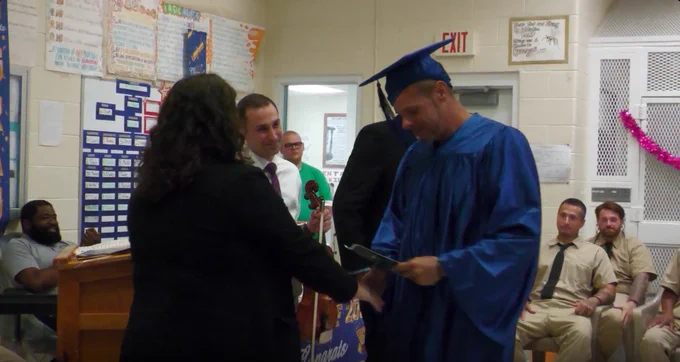 Graduate in blue cap and gown shaking hands with a woman during a ceremony in a classroom setting.