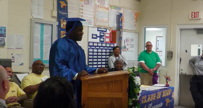 Graduate in blue cap and gown delivering a speech at a podium during a classroom graduation ceremony.