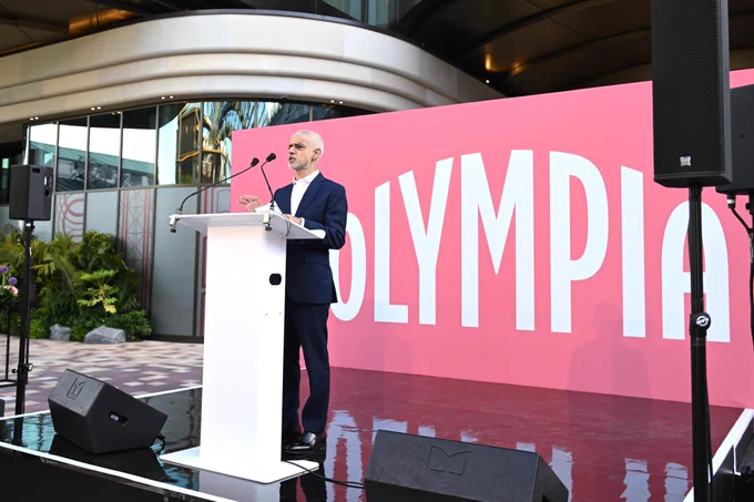 Mayor of London stood at a podium, pink backdrop displaying the words Olympia
