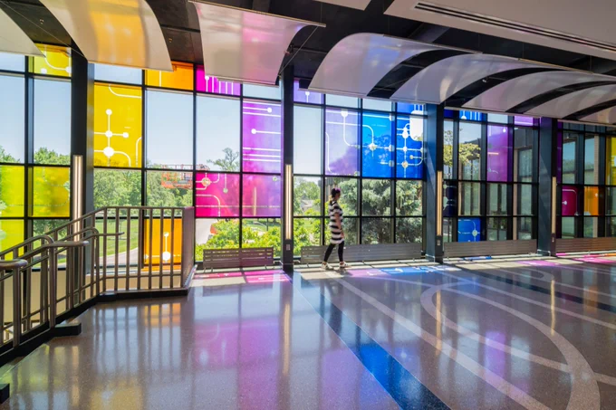 Sunlight streams through a colorful stained-glass installation in the College of Dentistry atrium, casting vibrant yellow, pink, purple and blue reflections across the floor. A person walks through the bright, open space, surrounded by large windows.