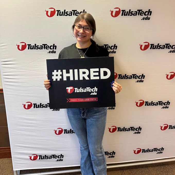 Person standing in front of a Tulsa Tech backdrop holding a sign that reads '#HIRED' with the Tulsa Tech logo and slogan.