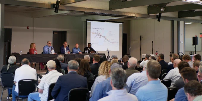 Five IBR speakers sit at a long table in front of a packed room full of construction industry professionals. 