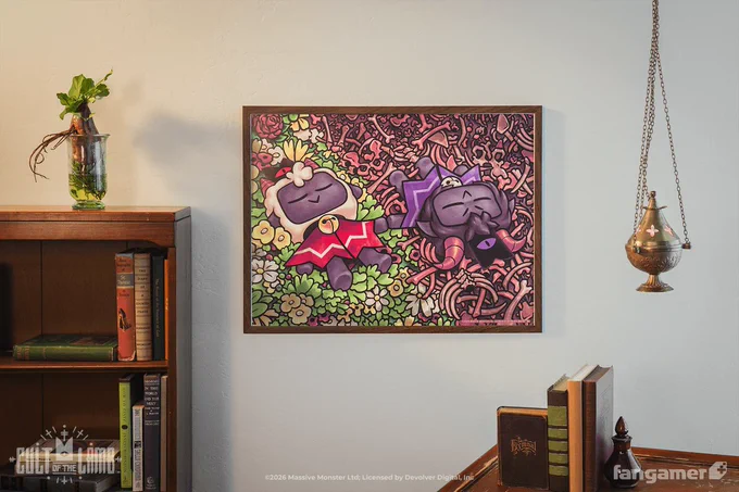 A photo of our new Cult of the Lamb poster framed on a wall, with a bookshelf agains the wall on one side and a table with books in the foreground on the other. It's a poster of the Lamb and the Goat lying peacefully in the grass (on the Lamb's side) or various bones (on the Goat's), with the illustration framed so that they're lying diagonally with a clean line down the middle where the landscape changes. 