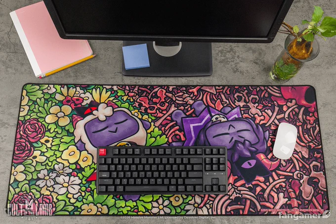 A photo of the same "Friends That Slay Together" illustration (with a more horizontal crop) on a desk mat, partially covered by a keyboard and mouse and arranged next to a notebook, a potted plant, and a notebook. 