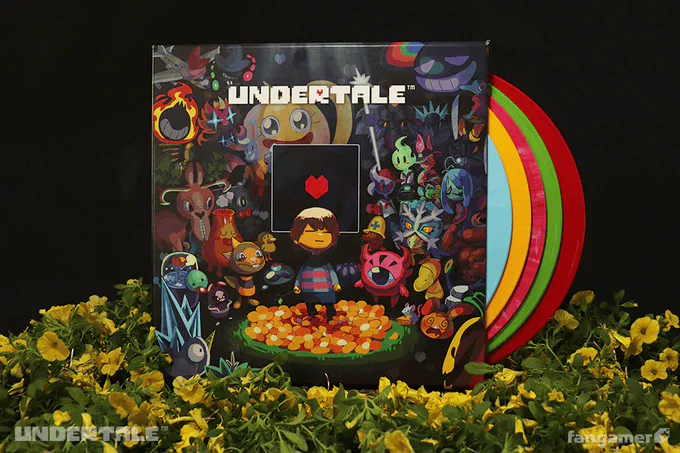 A photo of the UNDERTALE Complete Vinyl Soundtrack box set sitting on a bed of plastic plants and flowers in front of a black background. Five vinyl LPs are visible behind the packaging, which features a colorful illustration by ADE. 