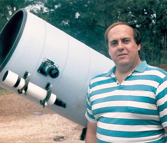 Photo of Alan Hale in a blue and white striped shirt with brown hair in front of a large white telescope