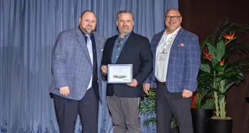Three men in suits stand together smiling, with one holding a framed award. They are on a stage with blue curtains and decorative plants, conveying a celebratory tone.