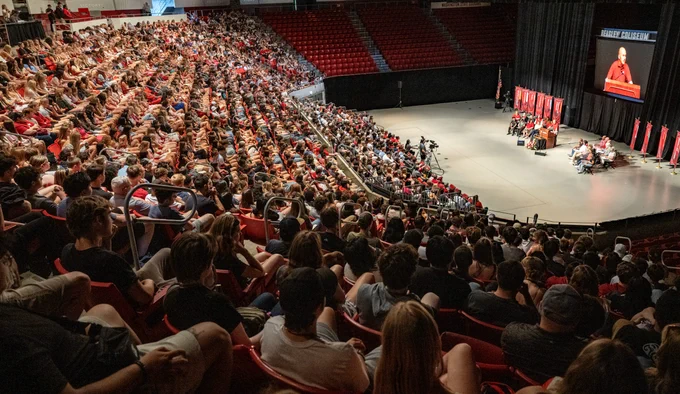 Wide-angle view of Beasley Coliseum filled with hundreds of seated students in red stadium-style seats, all facing a stage on the right. On the stage, several speakers sit behind a podium while one person speaks, shown on a large screen above with the text “Beasley Coliseum.” Red banners and an American flag stand near the stage, and cameras are set up in the aisle.