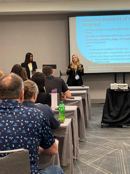 A white woman stands and holds her hands out gesturing while presenting to a room full of seated attendees. There is a Black woman watching from the side, standing at the presentation table. 