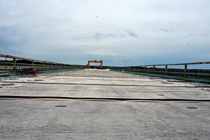 A concrete bridge stretches into the distance under an overcast sky. Metal guardrails line both sides of the structure, leading toward a piece of equipment or gate structure at the far end. Water is visible beyond the railings, and the scene appears to show infrastructure work at a dam, spillway, or navigation structure.