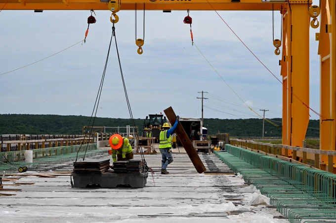 Two workers stand on a concrete bridge deck during maintenance work. One worker in a high-visibility safety vest and hard hat stands near equipment, while another works beside a sloped metal structure. An overhead crane with hanging hooks spans the top of the image. Guardrails line both sides of the bridge, and the cloudy sky creates an industrial worksite setting.