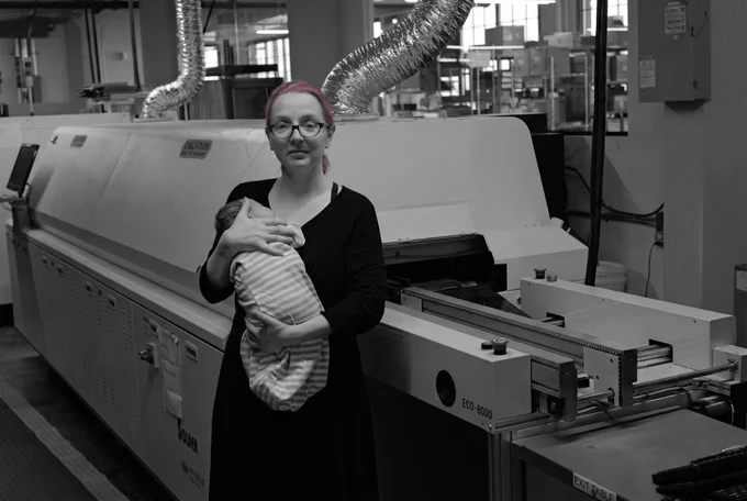 ladyada holding zine, her and pt's baby girl, in front of a reflow oven on the Adafruit factory floor in the USA, in BROOKLYN. black and white documentary photo, pink hair the only color in the frame. this is what's at stake when legal threats are used to silence security researchers and journalists - a woman owned engineering company in the usa, that has employees with families, people who report facts in good faith. stand up together.