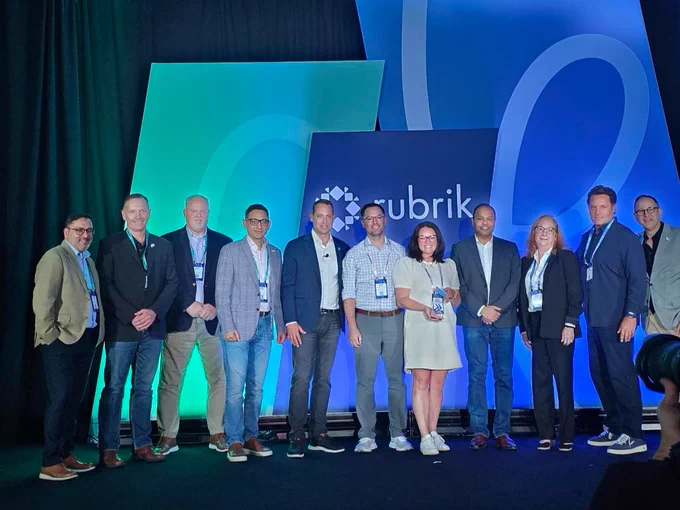 A group of individuals standing on a stage in front of a backdrop featuring the Rubrik logo. They are dressed in business attire, some wearing lanyards. The backdrop is illuminated with blue and green lighting.
