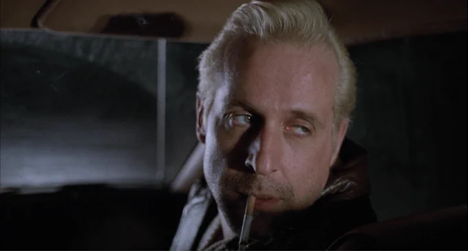 bleached blond Peter Stormare in closeup, looking bored as he looks behind him, cigarette dangling from his mouth 