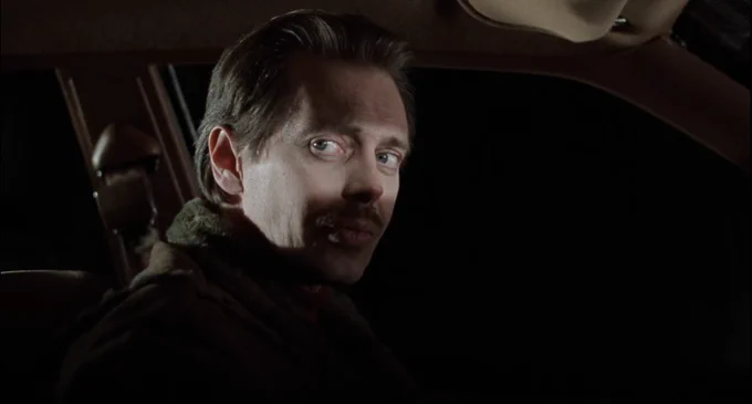 side view of Buscemi, his head partially turned toward the camera, his eyes and right cheek illuminated, a vast deep blackness visible beyond the driver's side window 