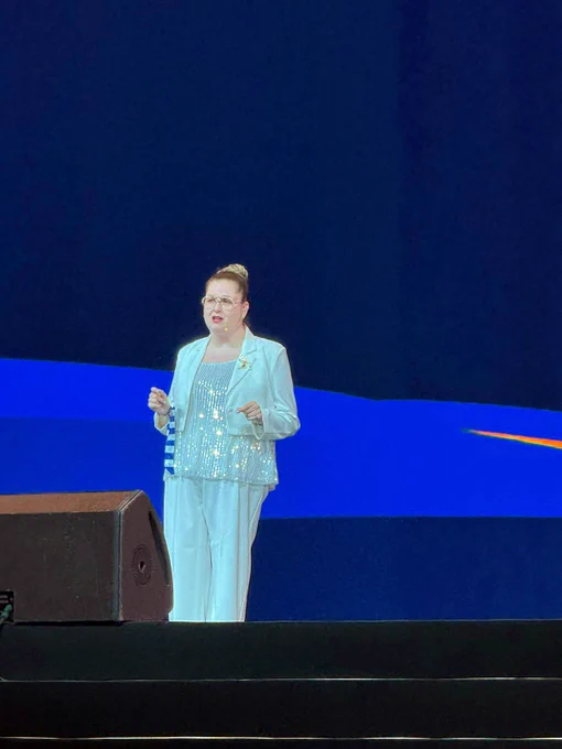 A person in a shimmering outfit stands on stage with a microphone, in front of a dark blue background with vibrant abstract shapes.