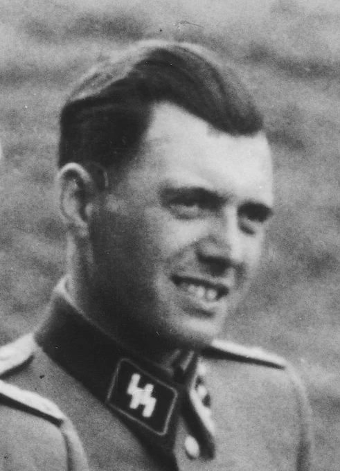 A black-and-white portrait of a man in an SS uniform - dr Josef Mengele. The uniform includes an SS insignia on the collar. The man has neatly combed hair and a slight smile