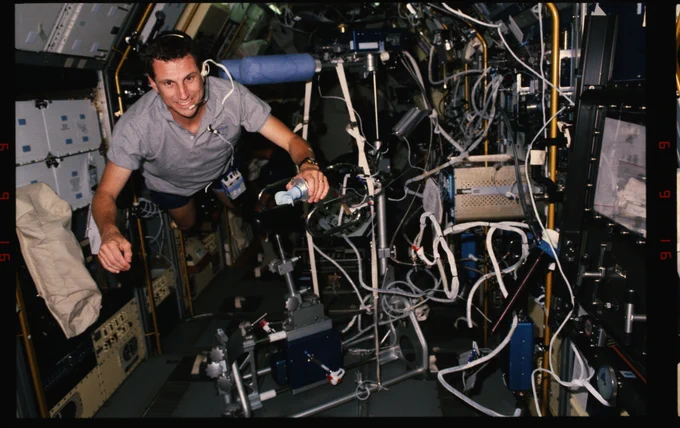 Wearing a gray short-sleeve shirt, F. Andrew Gaffney smiles while floating next to a bicycle ergometer and many cords and wires while aboard the space shuttle.