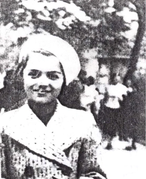 Old, grainy black and white photograph of a smiling individual in a coat, featuring a headscarf, standing in foreground with blurred figures in the background.