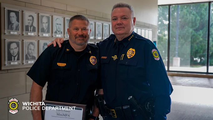 Two uniformed individuals from the Wichita Police Department pose together in a hallway, displaying framed portraits. One person holds a certificate. The department logo is visible in the corner.