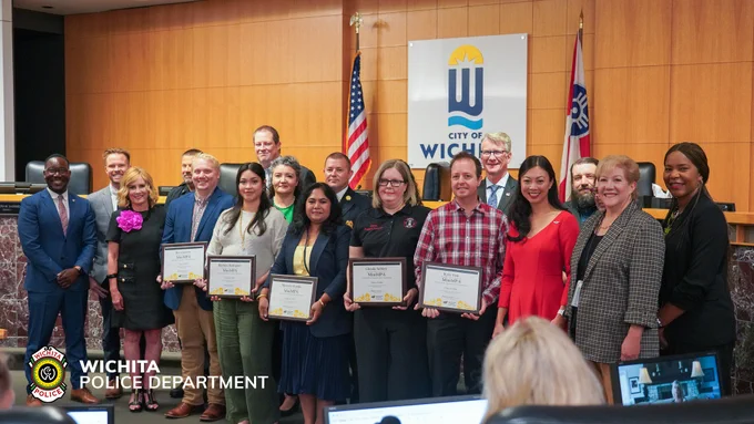 A group of individuals standing in a formal setting, some holding certificates. The Wichita Police Department logo is visible, and the City of Wichita emblem is displayed on the wall. Flags are positioned in the background.