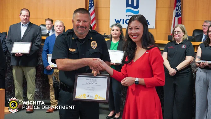 A group of people in a formal setting stand in front of a City of Wichita sign. One person in a police uniform shakes hands with another person in a red outfit, both smiling. Several others hold framed certificates. Wichita Police Department emblem is visible in the corner.