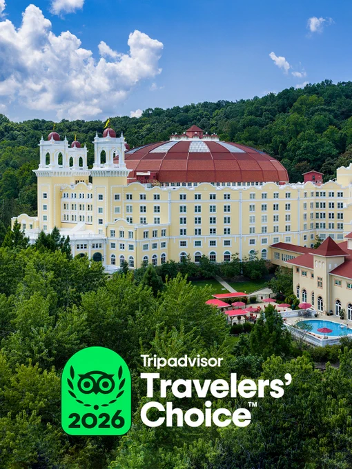 A large, West Baden Springs Hotel in southern Indiana surrounded by trees is displayed. The building has two towers with domed roofs and a spacious courtyard. The sky is partly cloudy, and the trees are lush green. In the foreground, a TripAdvisor Travelers’ Choice 2026 badge is visible.