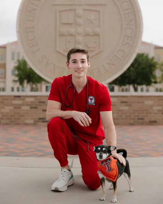 Male TTUHSC nursing student wears red scrubs and kneels down with his dog outside in front of the university seal.
