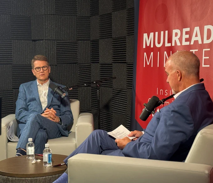 Two individuals are seated in a recording studio, engaged in a conversation. They are positioned opposite each other with microphones in front of them. A red banner in the background reads "MULREADY MINUTES." One person is holding a piece of paper. A small table with water bottles is in the foreground.
