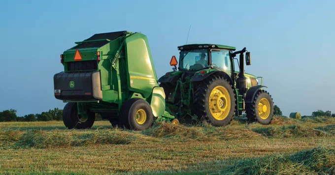 A John Deere tractor is attached to a green baler on a field of cut hay. The equipment is parked under a clear blue sky, with trees visible in the distant background.