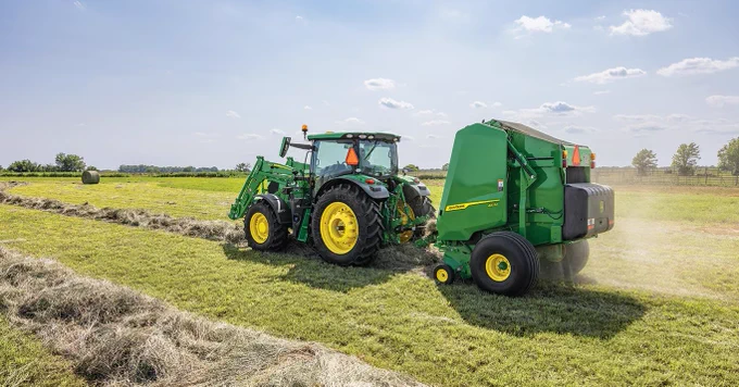 A green John Deere tractor is pulling a large green baler across a grassy field, forming cylindrical bales of hay. The sky is clear with some clouds.