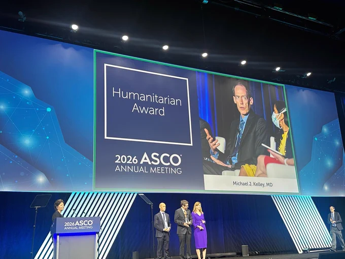 The image shows a stage at the 2026 ASCO Annual Meeting featuring a large screen displaying "Humanitarian Award" and an image of a speaker with the name Michael J. Kelley, MD. On stage, there are four individuals, and one stands at a podium with the ASCO logo. The setting is formal, with blue lighting and a professional atmosphere.