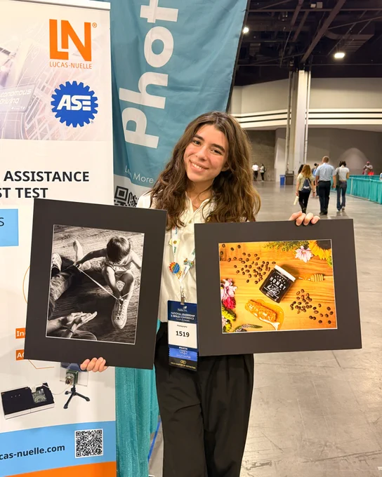 Person holding two framed photographs at an indoor event, one black-and-white of a child drawing and one colorful with coffee beans and a cup.