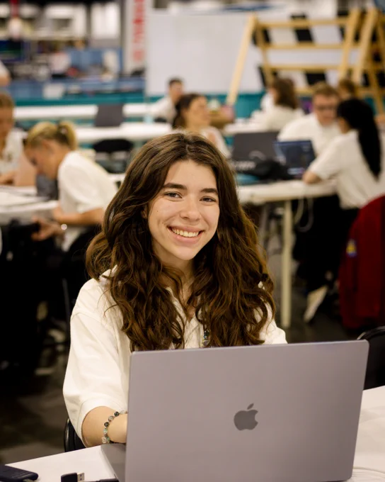 Young woman with long curly hair working on a silver Apple laptop in a busy office environment with colleagues in the background.