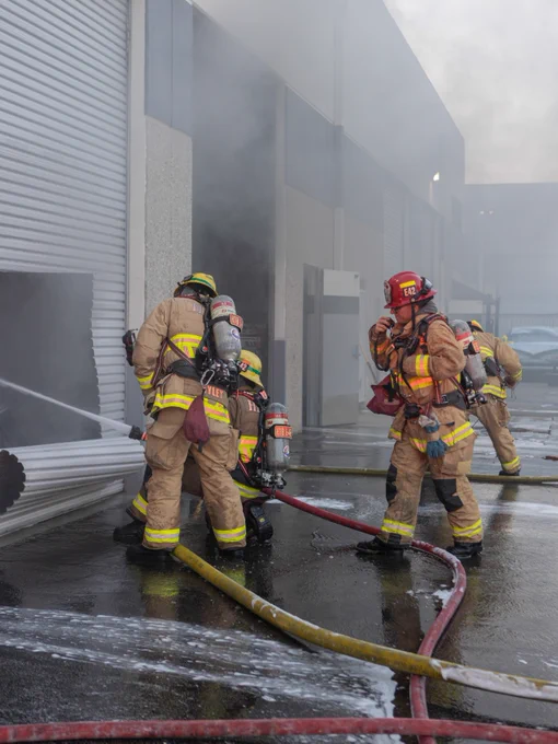 Firefighters operate outside a commercial warehouse unit as smoke vents from an open roll up door. One crew advances a hose line into the structure while additional firefighters coordinate operations in the smoky industrial complex.
