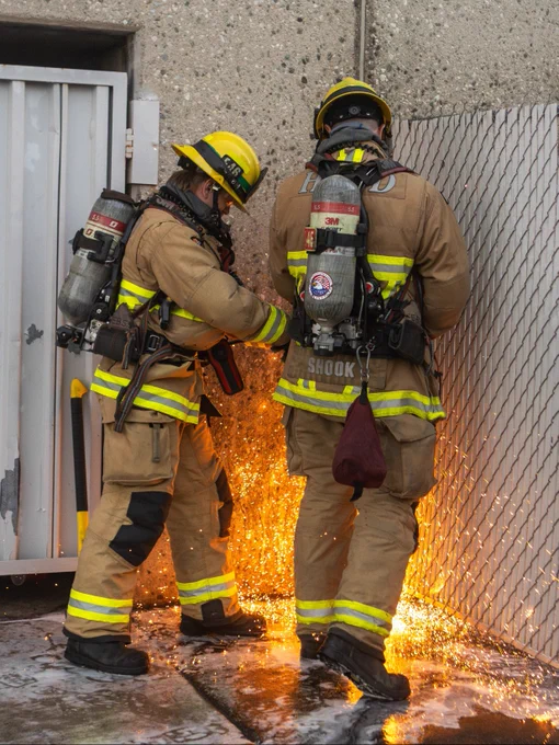 Two Huntington Beach firefighters in full protective gear use a rotary saw to perform forcible entry on a metal security gate at a commercial building. Bright sparks shower across the ground as crews work to gain access to the fire area.