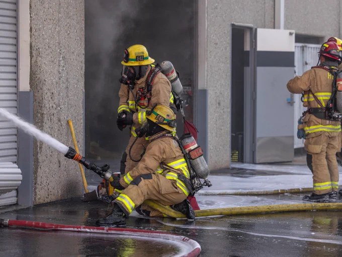 Firefighters direct a hose stream through a partially opened roll up door of a commercial warehouse while heavy smoke escapes from the interior. Additional personnel monitor conditions and communicate tactical assignments nearby.