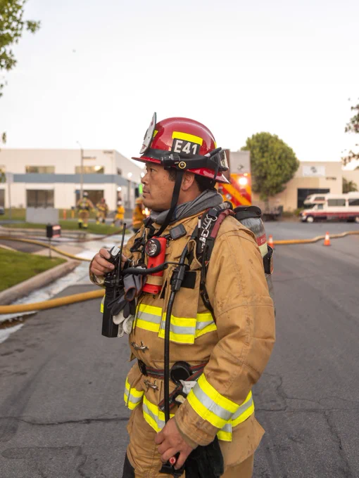 Captain Keita Sato of the Huntington Beach Fire Department stands in the street with a portable radio during commercial structure fire operations. Fire apparatus, hose lines, and firefighters are visible in the background as incident command activities continue.