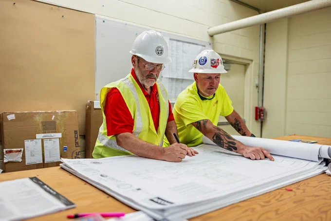 Two Division of Industrial Compliance inspectors wearing safety vests and helmets review large architectural plans on a table.