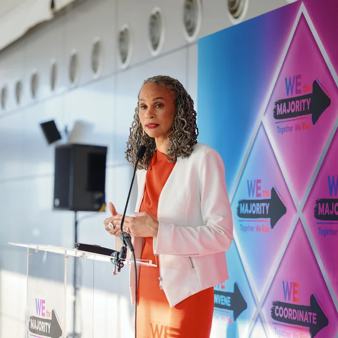 LCCHR President and CEO Maya Wiley speaking in front of a backdrop with the text "We the Majority" and "Together We Rise." Wiley is wearing an orange outfit with a white blazer.