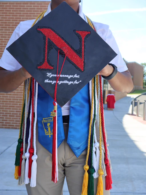 A person holds a black graduation cap with a red/black beaded 'N' and white text: \"If you wanna go far, then you gotta go far\". They wear numerous colorful honor cords & a blue \"National Honor Society\" stole. A red/white \"2024\" tassel hangs from the cap.