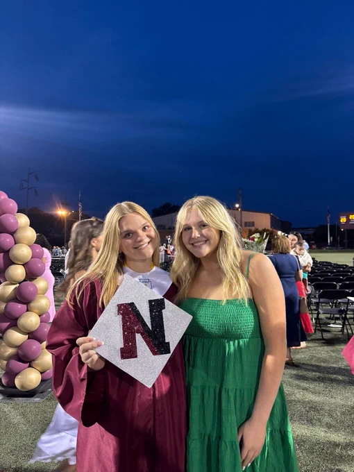Two smiling young women with blonde hair stand outdoors at dusk. One in a maroon graduation gown holds a glittery silver graduation cap with a large red and black 'N'. The other wears a green tiered dress. In the background are empty black chairs and a scoreboard.