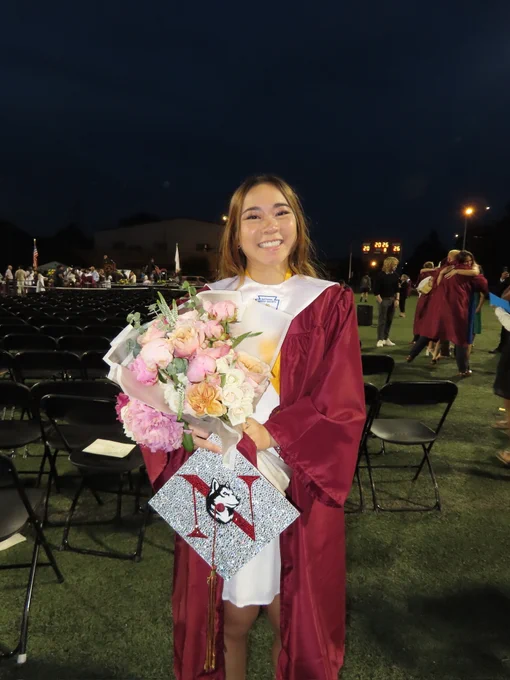 Smiling graduate in a maroon gown and white stole holds a bouquet of pink, peach, and white flowers. Her silver glitter graduation cap has a red 'X' and a husky logo. Behind her, empty chairs on a green field at a nighttime ceremony.
